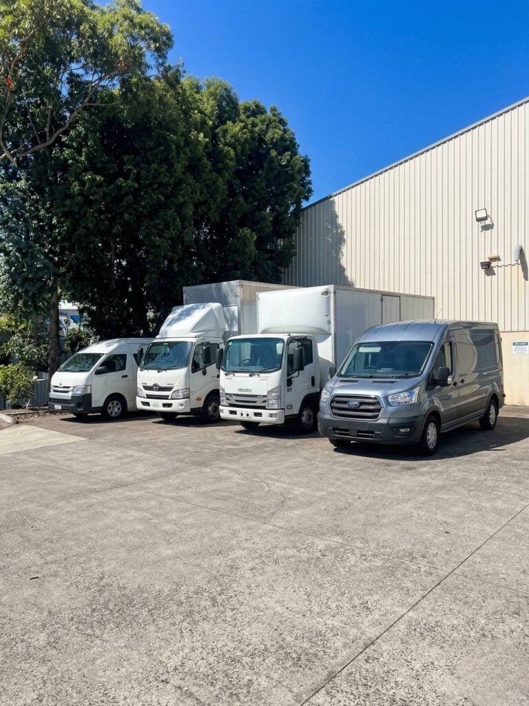 A grey Ford cargo van, two white box trucks, and a white Toyota van parked in a line outside a warehouse under a bright blue sky.