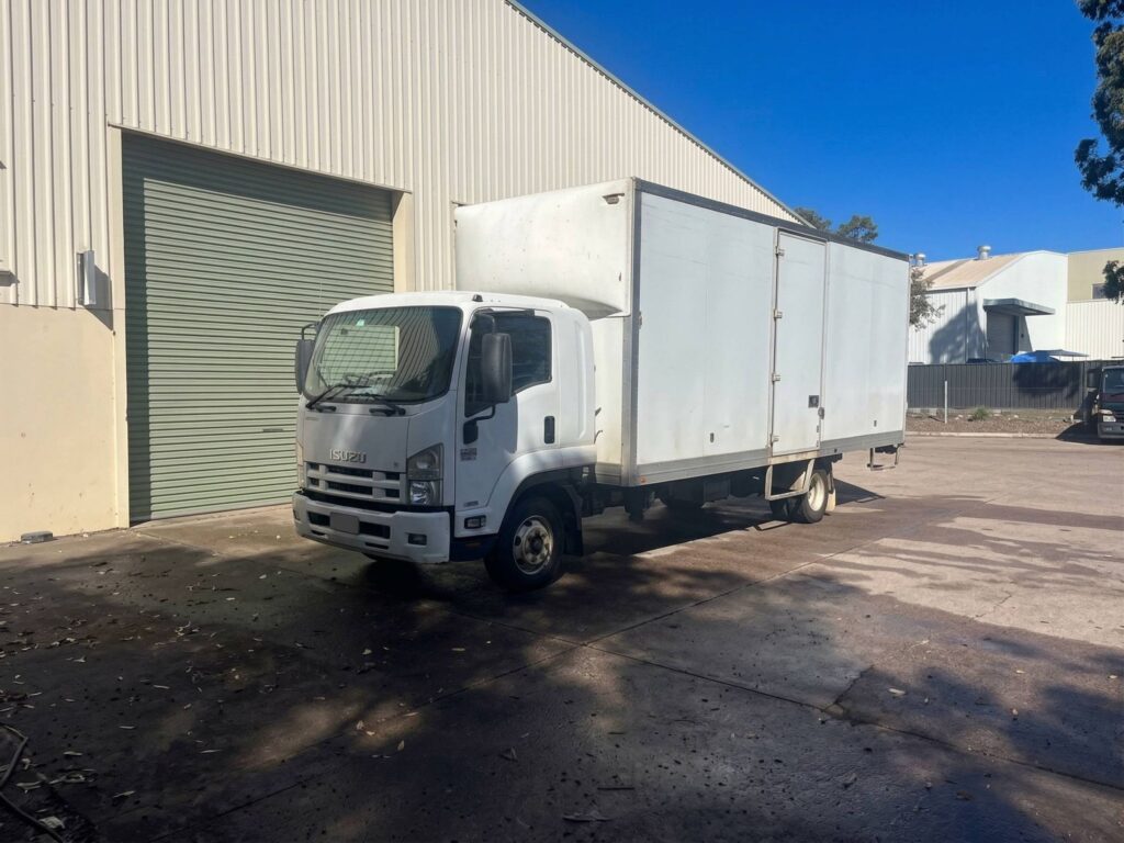 A white Isuzu box truck parked on a concrete lot next to a warehouse building with a large roller door under a clear blue sky.